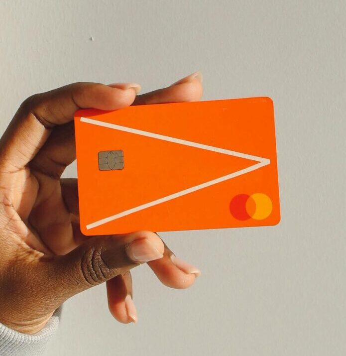 Close-up of a hand holding a bright orange debit card against a light background.